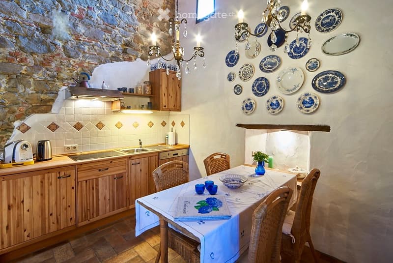 Cozy kitchen with wooden cabinets, stone wall, chandelier, and decorative plates. Includes a dining table with chairs, cups, and a bowl.