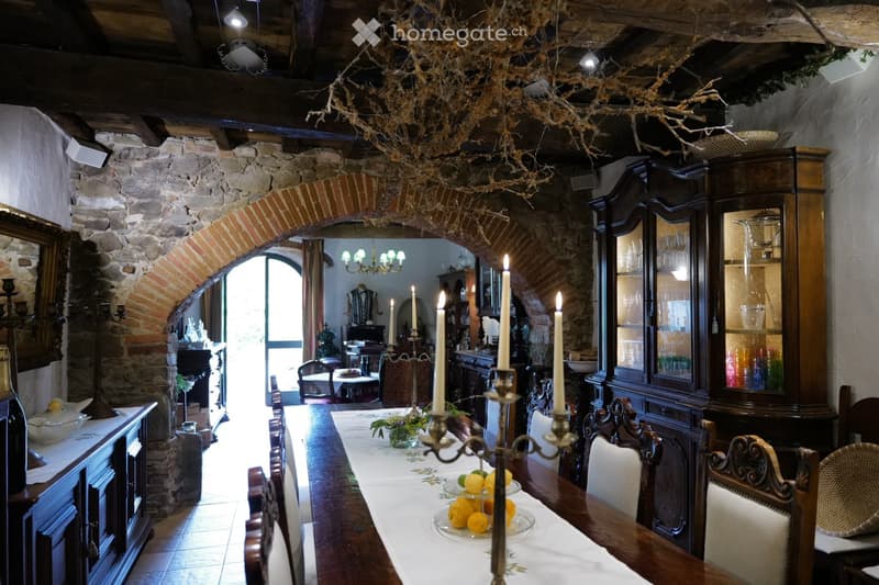 Elegant dining room with exposed wooden beams, stone walls, and a long wooden dining table set with white tablecloth, fruit bowl, and candle holders. Ornate wooden chairs and cabinets line the room, with an arched brick opening leading to another room.