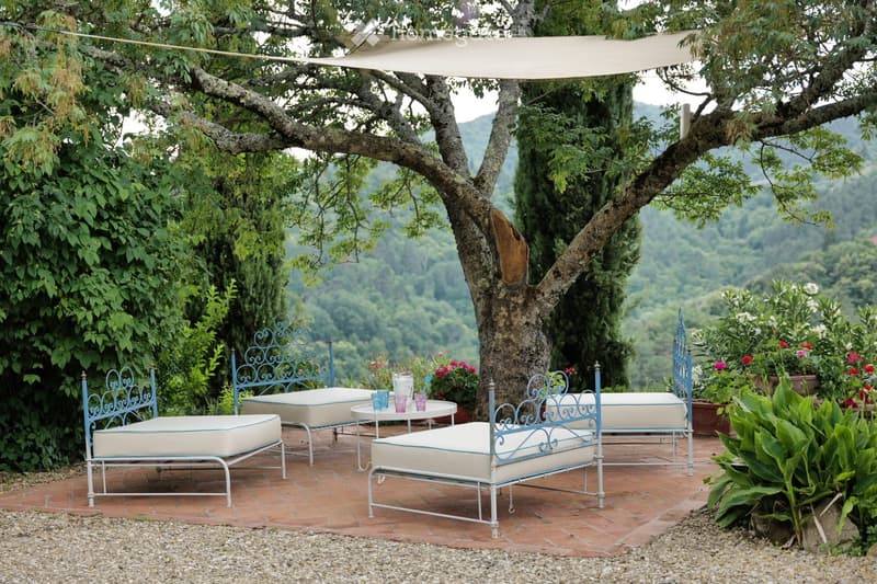A serene outdoor seating area with a large tree providing shade, featuring multiple white cushioned benches with blue metal frames, a small round table with cups, and lush greenery in the background.