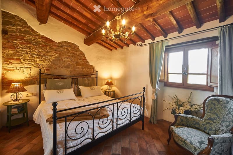 Cozy bedroom with exposed wooden beams and stone walls, featuring a metal-framed bed, nightstands with lamps, a patterned armchair, and a chandelier.