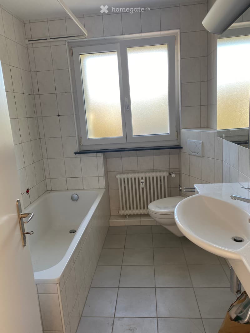 Bathroom with a bathtub, toilet, sink, and radiator. The room features tiled walls and floor, a window with frosted glass, and a door with a gold handle.
