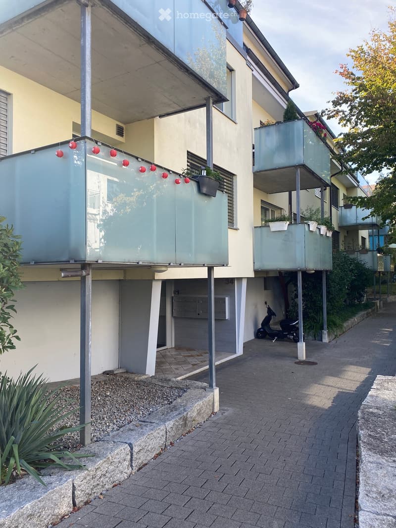 Modern multi-story building with glass balconies, light-colored facade, and decorative hanging lights. Features a paved walkway and small garden area with plants.
