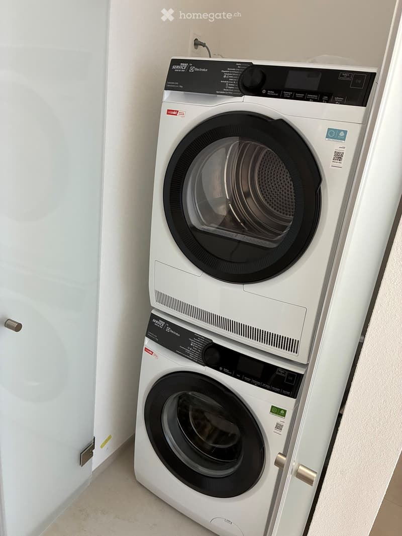 A modern bathroom with a white washing machine and dryer stacked vertically, featuring a sleek design and control panels.