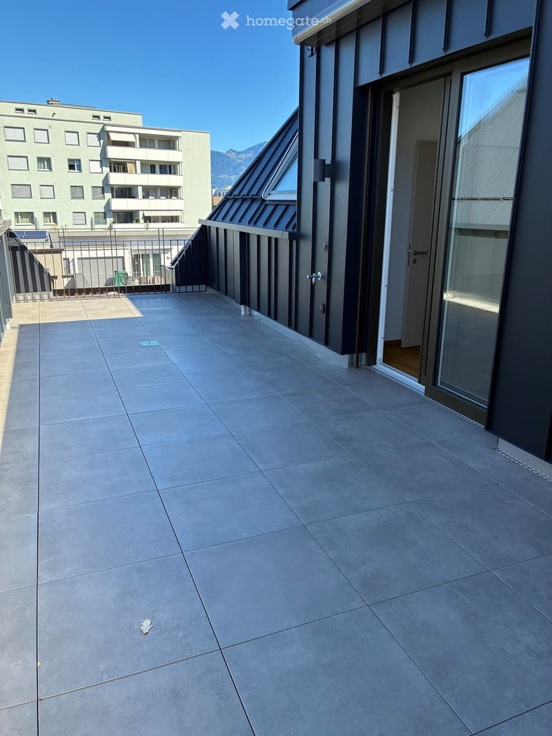 Modern balcony with concrete tiles, glass door, metal railing, and a view of neighboring buildings and mountains.
