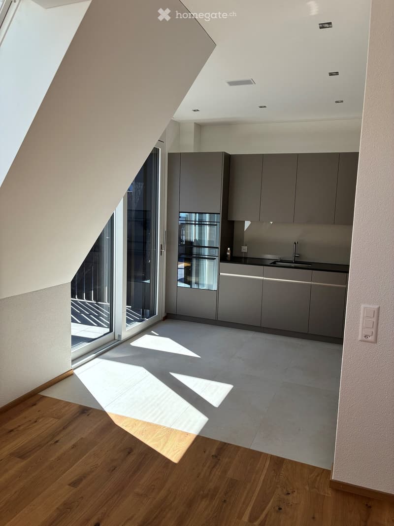 Modern, minimalist kitchen area with grey cabinets, stainless steel appliances, and a large window letting in natural light. The space is unfurnished, showing only the built-in cabinetry and appliances.