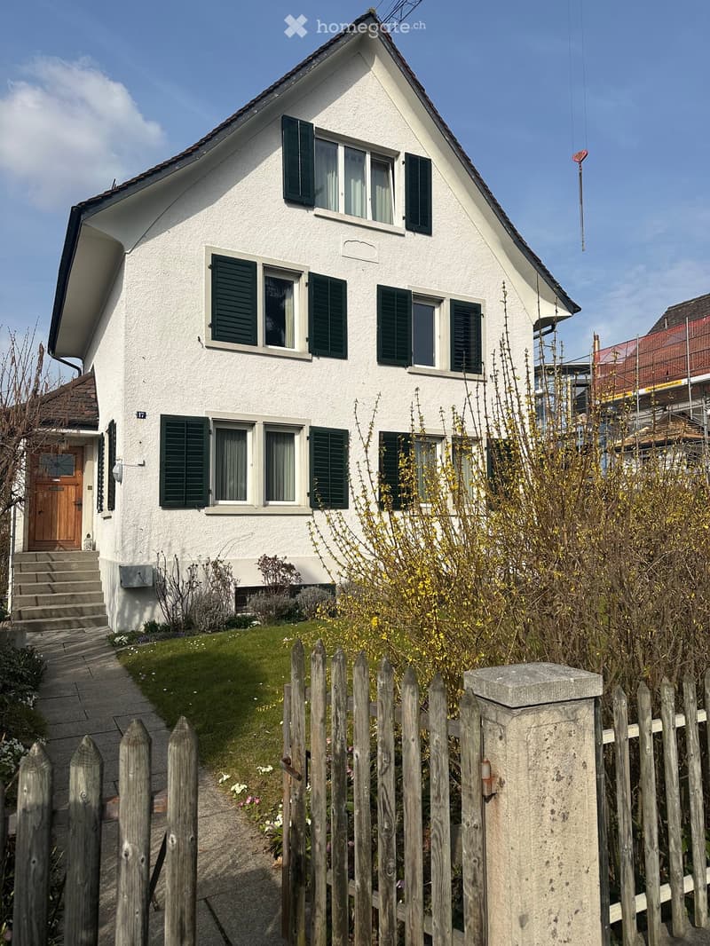 Two-story white house with green shutters, wooden front door, and small garden behind a wooden fence.