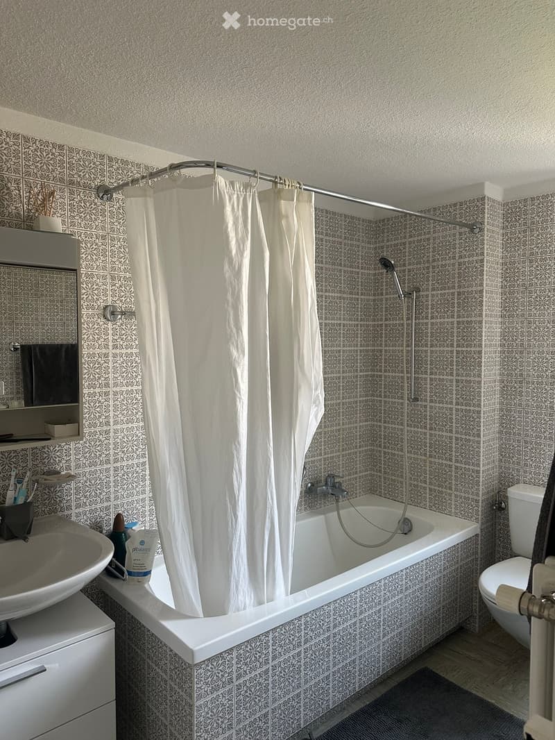 Bathroom with a bathtub, toilet, sink, and shower curtain. The walls are tiled with a patterned design, and there is a mirror above the sink.