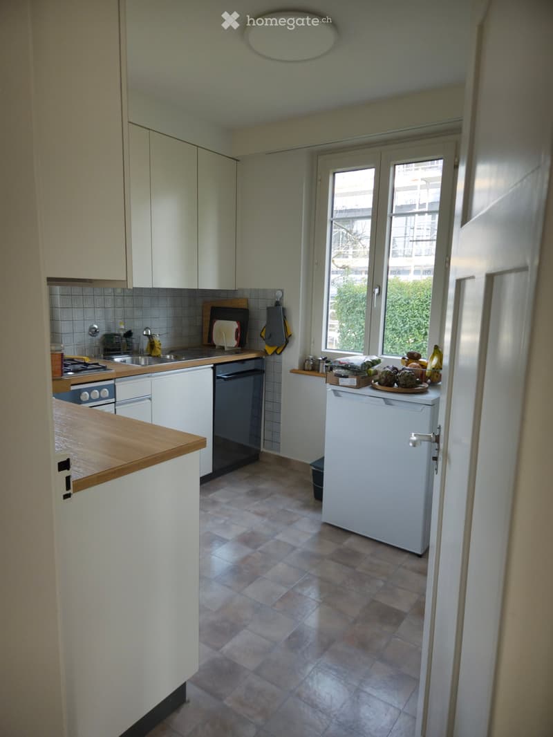 The kitchen features white cabinets, a wooden countertop, a tiled backsplash, and a tiled floor. Visible appliances include a refrigerator, oven, and sink. A cutting board and oven mitt are placed on the counter, along with a dish rack and a few kitchen i