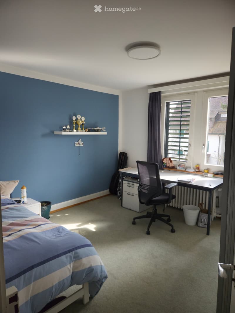 Bedroom with a bed, desk, chair, shelf with trophies, guitar, and window with curtains.