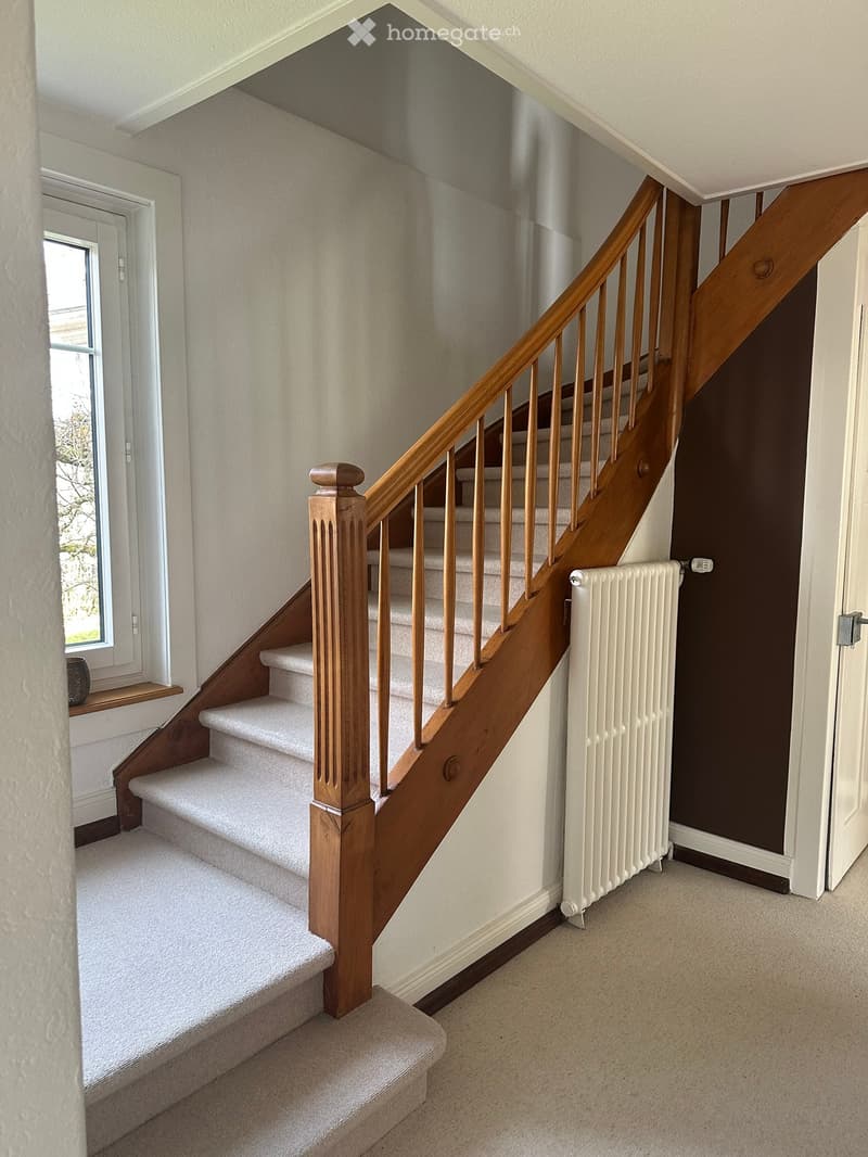 Spacious hallway with a wooden staircase featuring a polished handrail and balusters, carpeted stairs, a white radiator, and a window letting in natural light.
