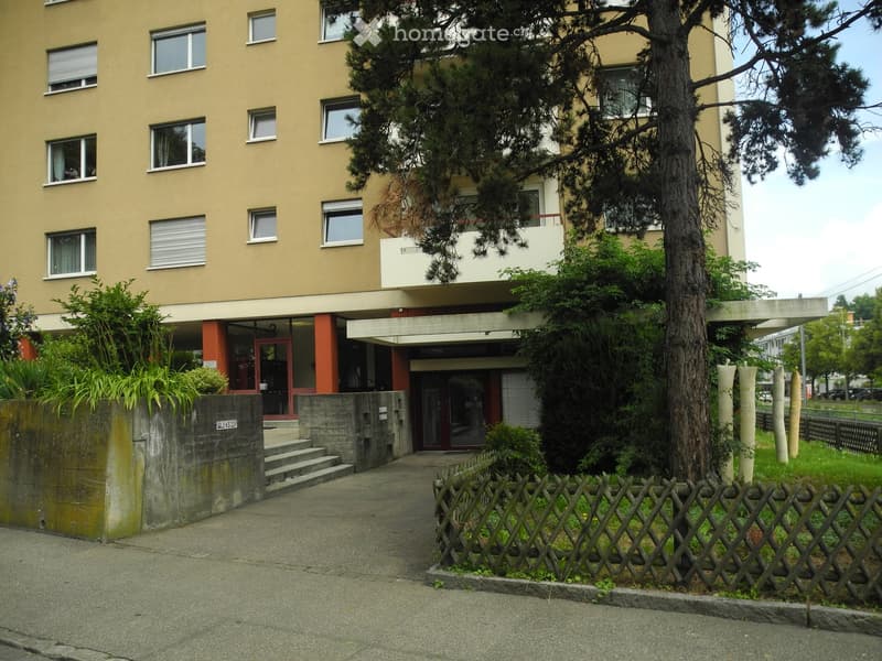 Multi-story residential building with beige facade, multiple windows, and a tree in front. The entrance features a small staircase and a red door.