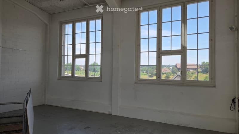 Empty room with bare concrete walls and floor, large windows offering a view of the surrounding area.