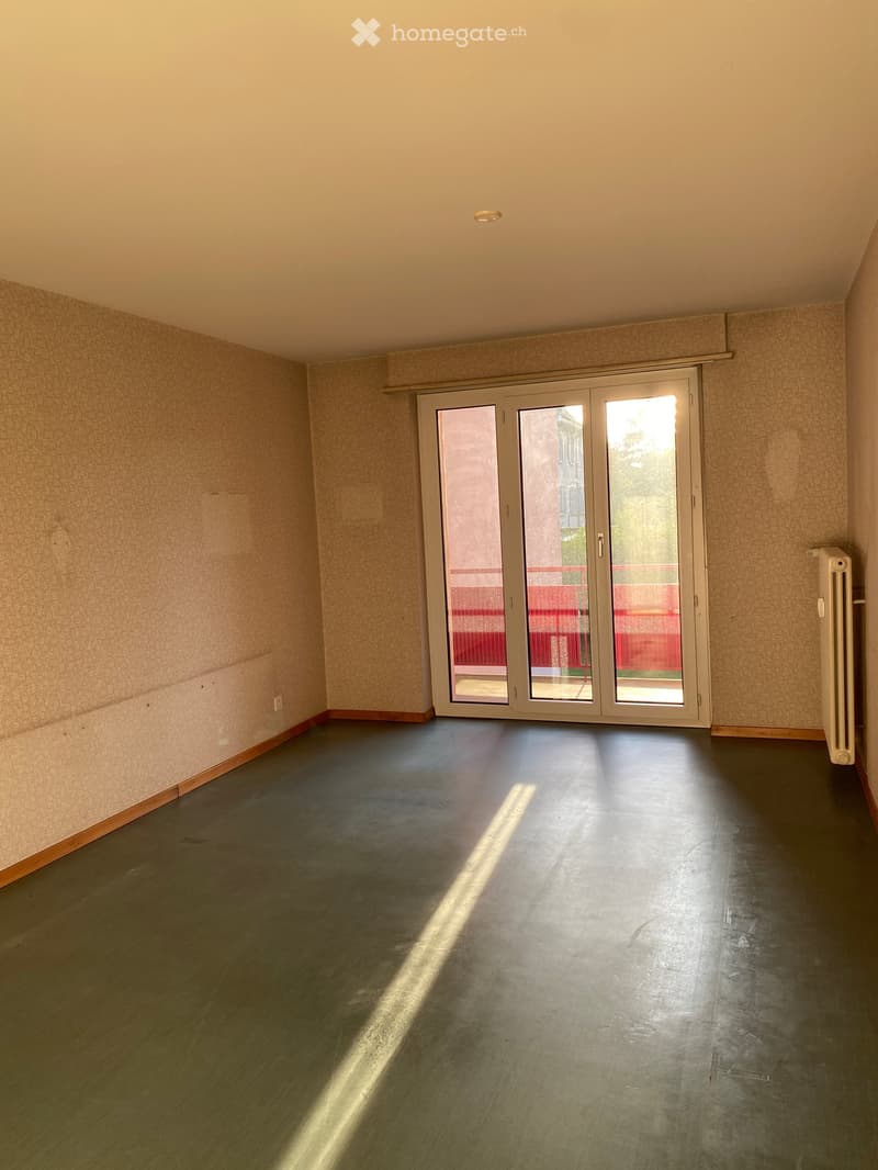 Empty room with grey concrete floor, beige walls, large sliding glass doors leading to a balcony, and a white radiator on the right side.