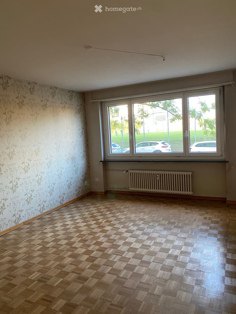 Empty room with wooden parquet flooring, floral wallpaper on one wall, large windows with blinds, radiator under the windows, and visible electrical outlets.