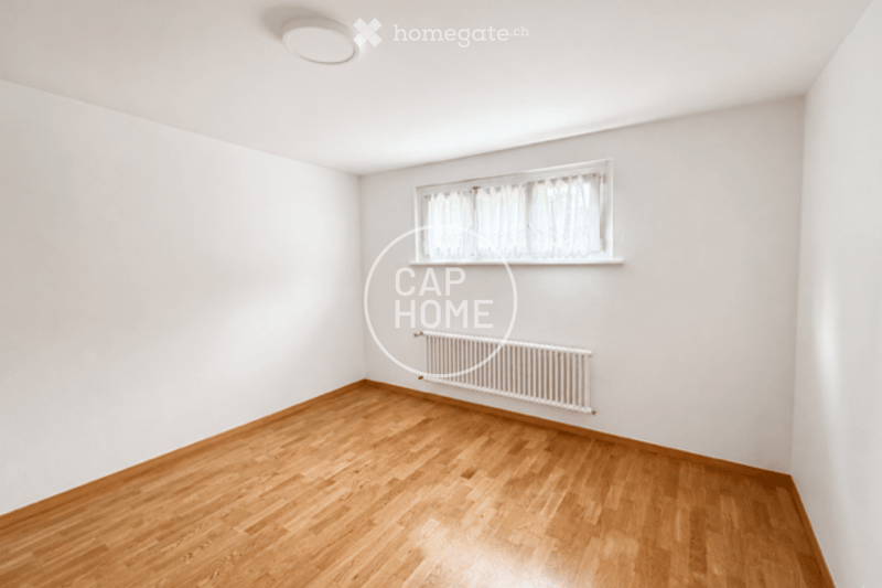 Empty room with wooden flooring, white walls, a window with white curtains, and a radiator below the window.