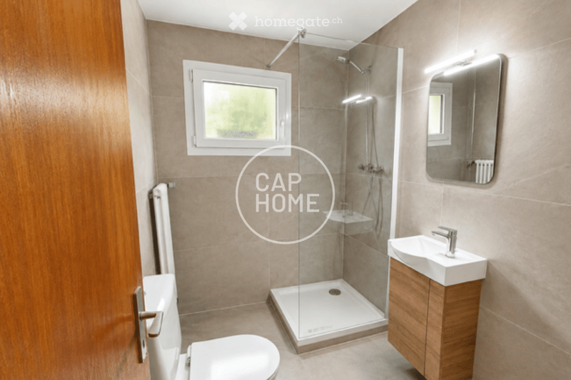 Modern bathroom with a glass-enclosed shower, a white toilet, a wooden vanity with a sink, and a large mirror above the sink. The walls are tiled in a neutral color, and there is a window providing natural light.