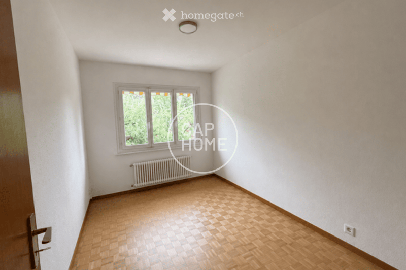 Empty room with wooden flooring, white walls, a window with multiple panes, and a radiator below the window. The room has a simple, clean design with no furniture or decorations.