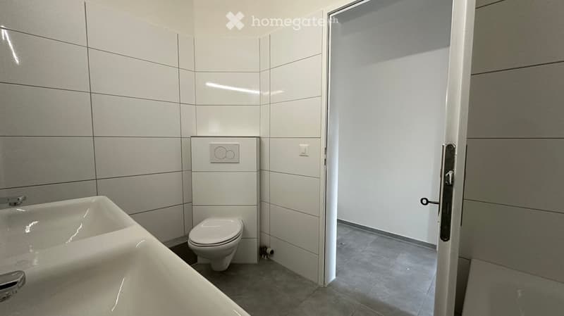 Modern bathroom with white tiles, a bathtub, and a toilet. The door is open, revealing a clean and contemporary design.
