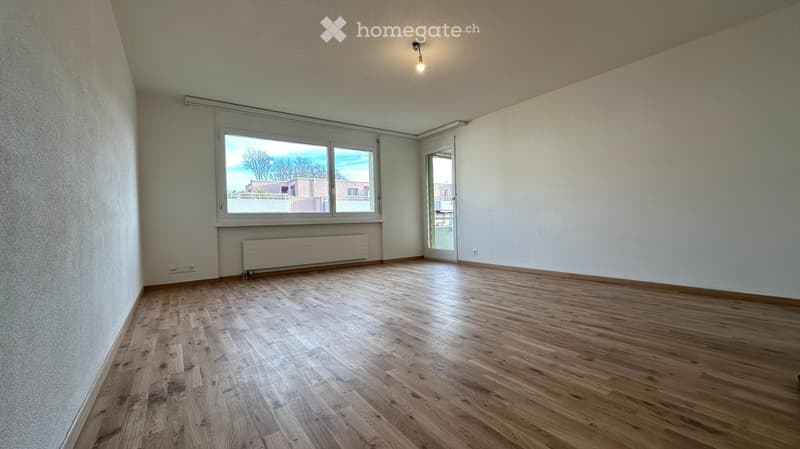Empty room with wooden flooring, large window, radiator, and a sliding door leading to another space.