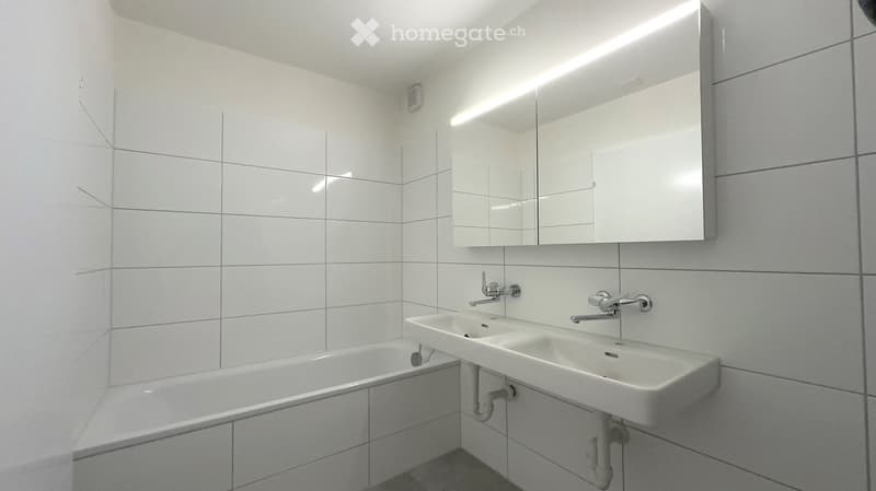 Modern bathroom with white tiled walls, a bathtub, a double sink, and a large mirrored cabinet with built-in lighting.