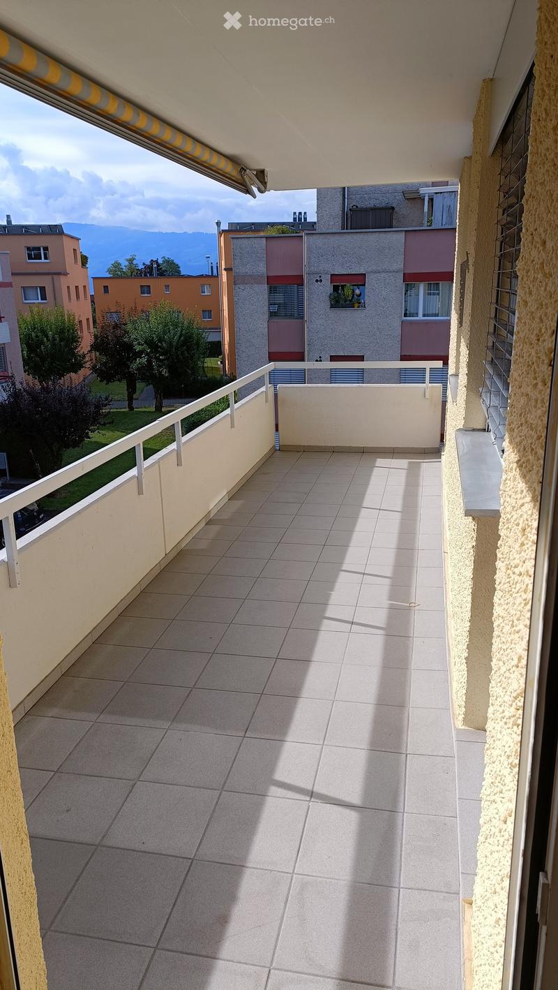 Balcony with tiled floor, white railing, and a view of surrounding buildings and greenery.