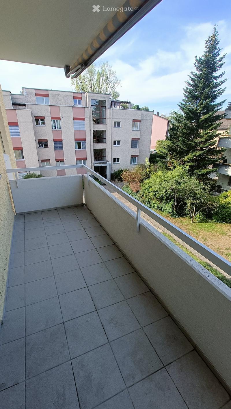 Balcony with tiled floor, white railing, and view of surrounding buildings and greenery.