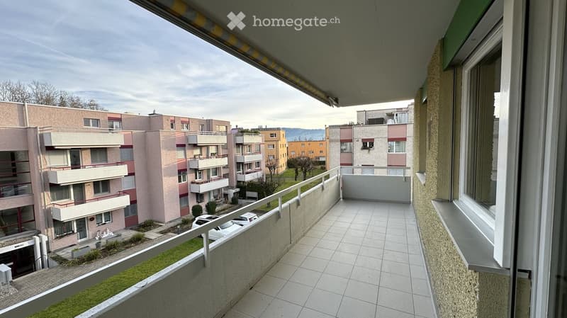 Balcony with tiled floor, metal railing, overlooking a residential area with multiple apartment buildings, greenery, and parked cars.