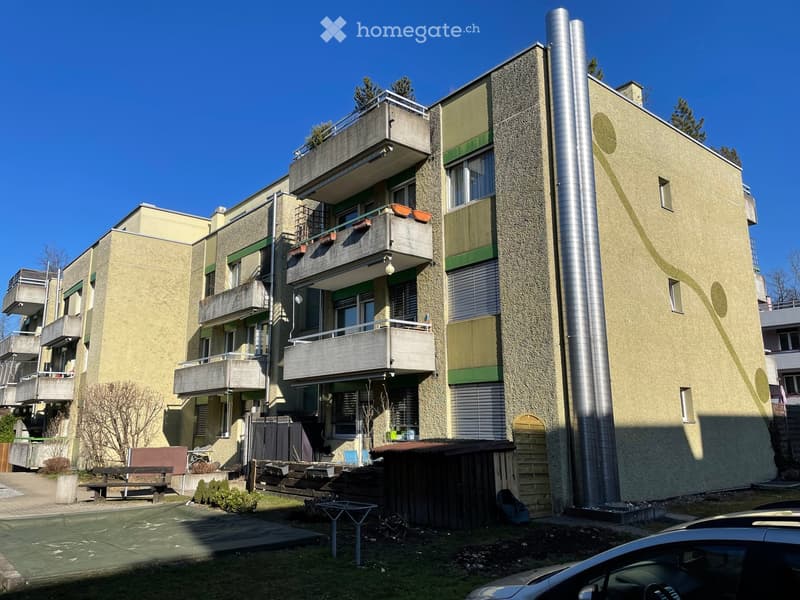 Modern multi-story residential building with balconies, green accents, and a small outdoor area with benches and a table.