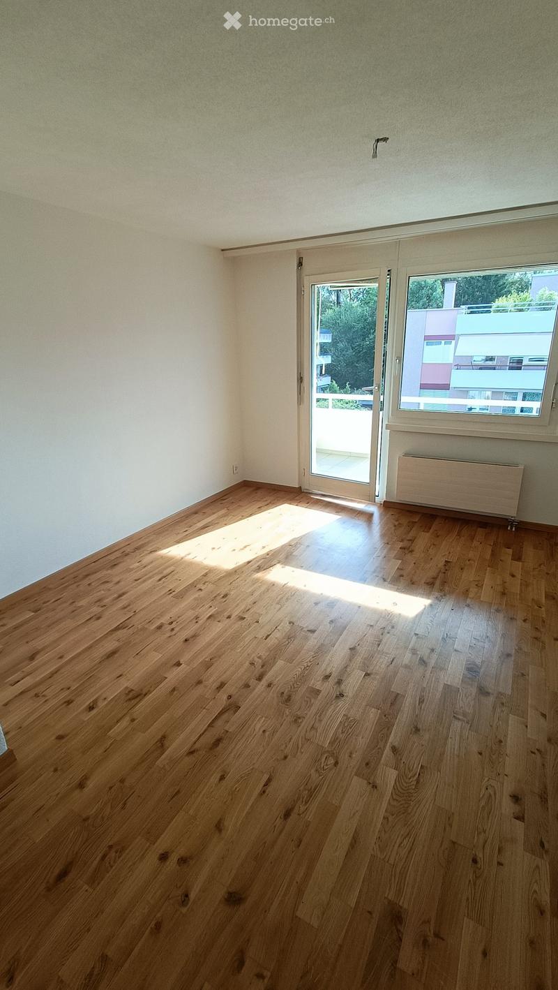 Empty room with wooden flooring, white walls, large window, and sliding glass door leading to a balcony.