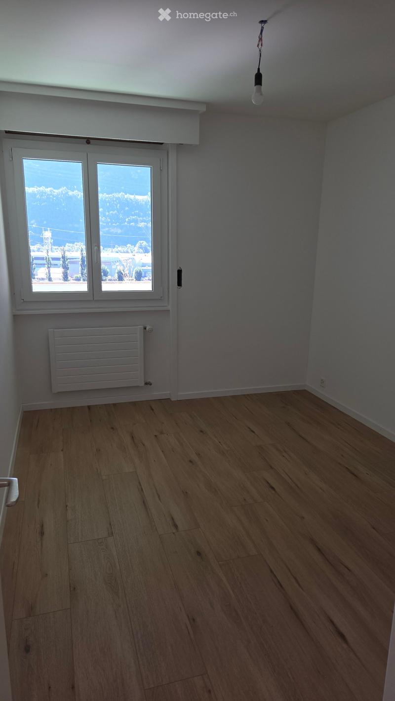 Empty room with wooden flooring, white walls, a window with a view, a radiator, and a single hanging light fixture.
