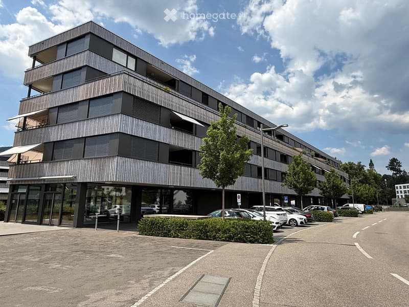 Modern multi-story building with balconies, large windows, and a parking area with several cars.