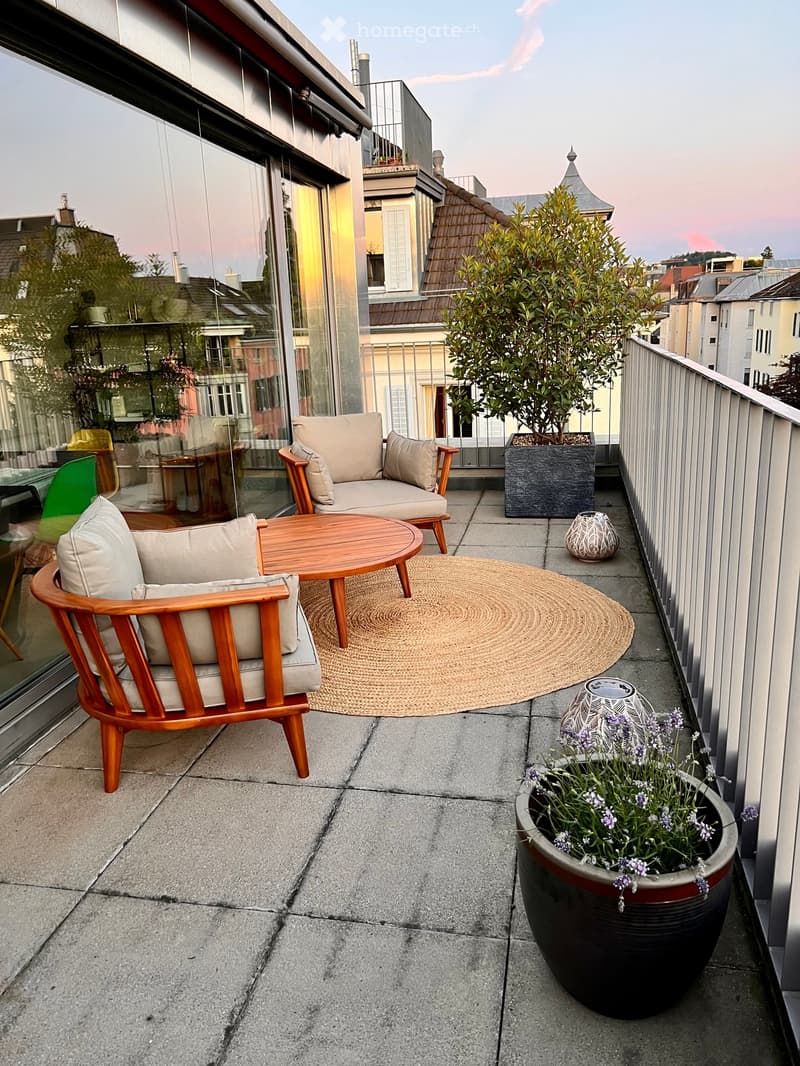 Balcony with two wooden chairs, a small round table, a woven rug, potted plants, and decorative vases.