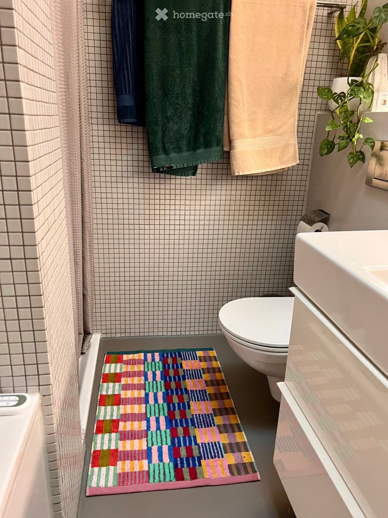 Modern bathroom with white tiles, a colorful bath mat, a toilet, a sink, and a small plant on the edge of the sink.