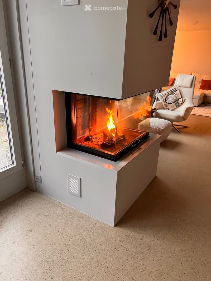 Modern living room with a sleek white fireplace, a white sofa, a white armchair, and an ottoman. The fireplace has a glass enclosure with a visible flame.