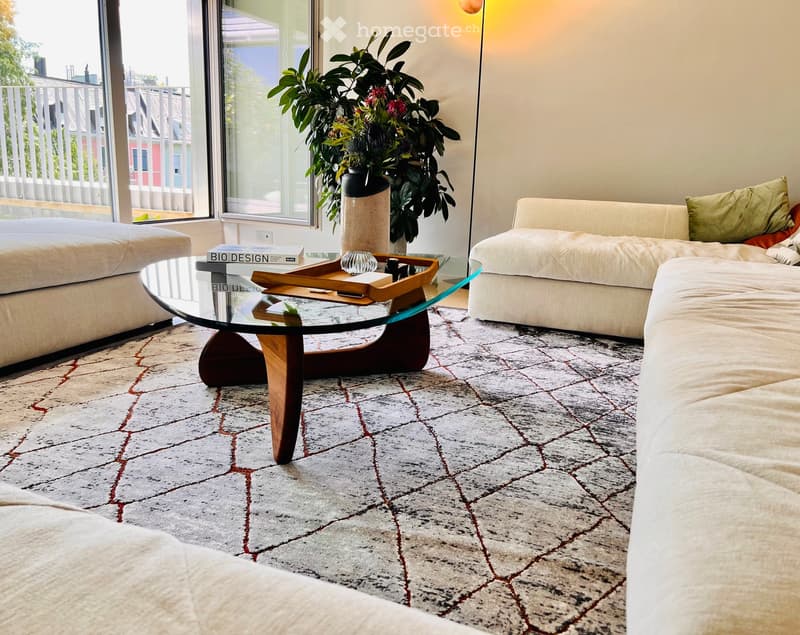 Modern living room with a large window offering a view, featuring a white sectional sofa, a glass coffee table with wooden base, an ottoman, a large plant in a pot, and books on the coffee table.