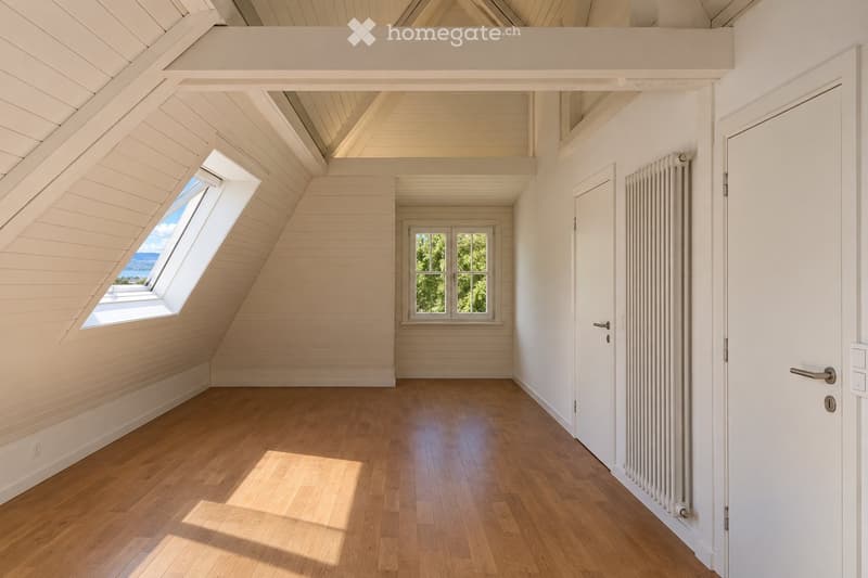 Bright, empty attic room with wooden floors, white paneled walls, and large windows offering natural light and a view of greenery.