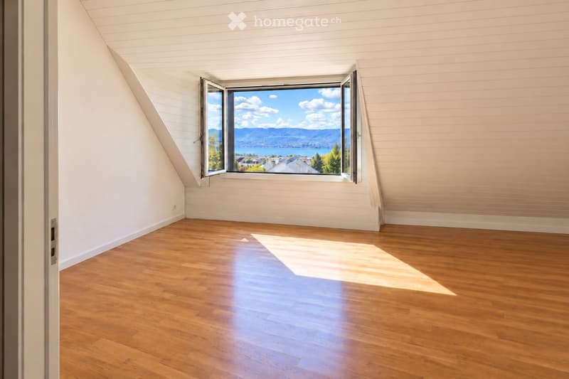 Empty room with wooden flooring and white walls, featuring a large window offering a scenic view of a lake and mountains.