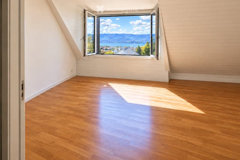 Empty room with wooden flooring, white walls, and large windows offering a scenic view of a lake and mountains.
