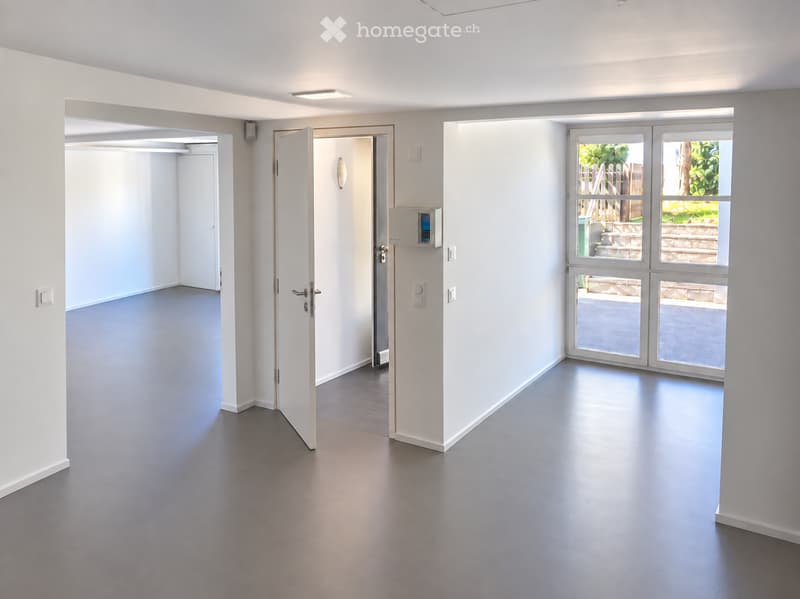 Empty room with white walls, grey flooring, and large windows leading to an outdoor area. No furniture or equipment is present.