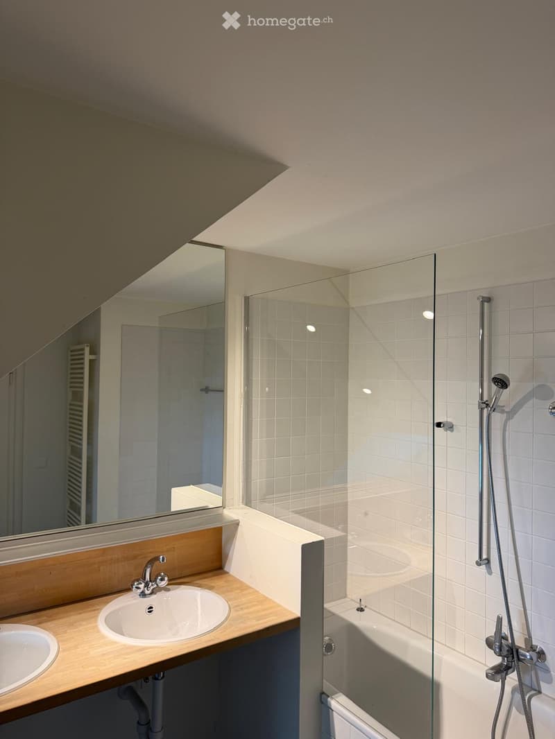 Modern bathroom with a wooden vanity, two sinks, a large mirror, a bathtub with a glass shower enclosure, and a handheld showerhead.
