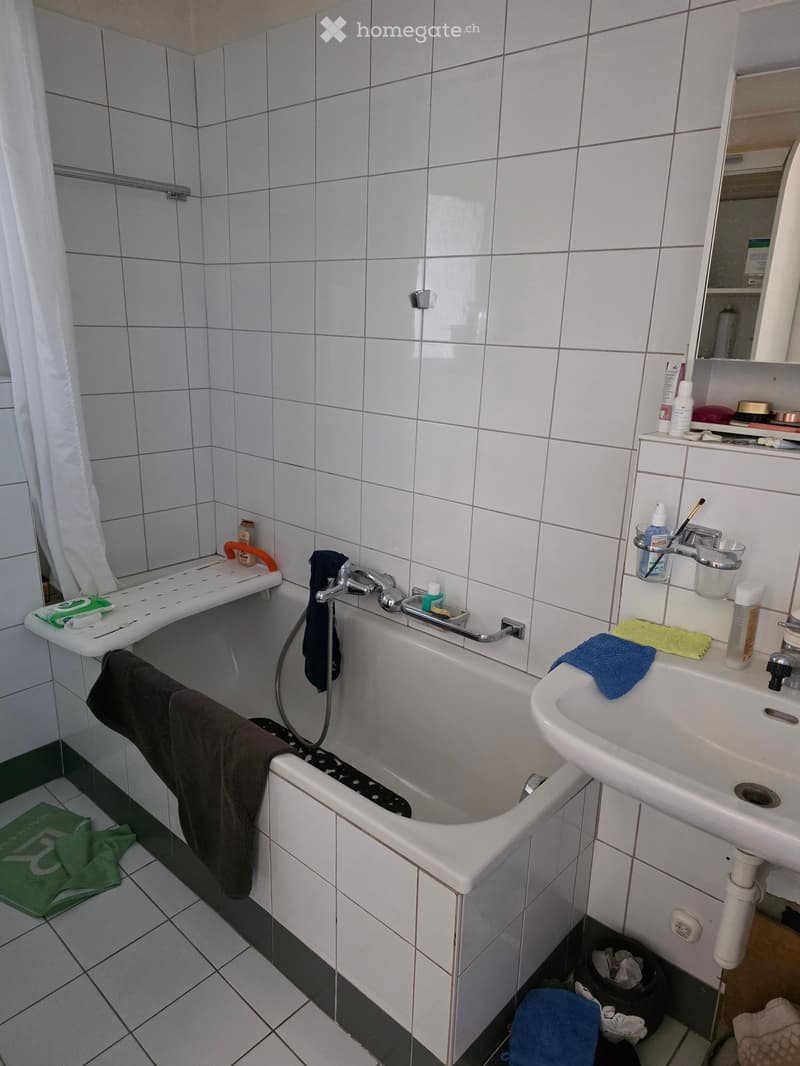 Bathroom with white tiled walls and floor, bathtub with handrail, sink with mirror, various toiletries, and towels.