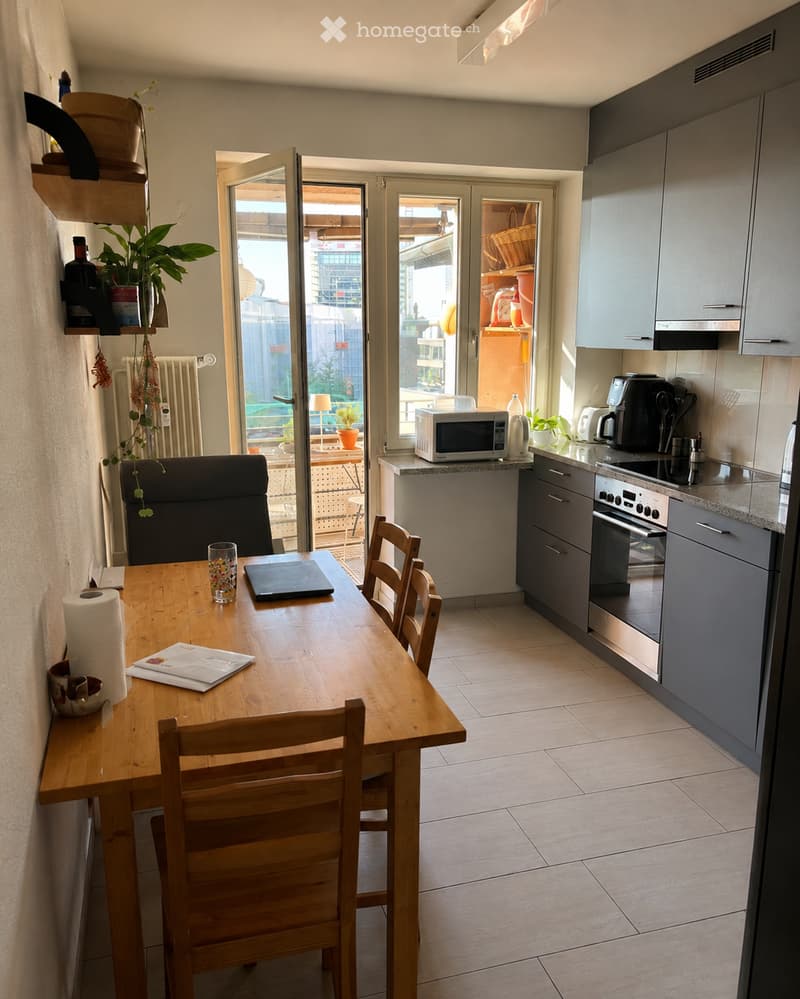 A modern kitchen and dining area with a wooden dining table, four chairs, a laptop, a microwave, an oven, and a coffee maker. The space features grey cabinets, a tiled floor, and a large window with a view of the city.