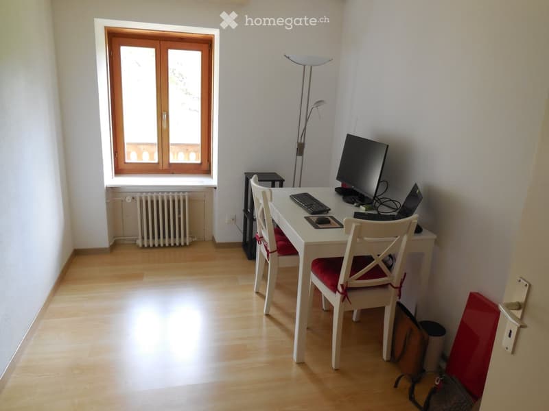 Bright, furnished room with wooden flooring, a white desk, two chairs with red cushions, a laptop, keyboard, mouse, and a floor lamp near a large window with wooden frames.