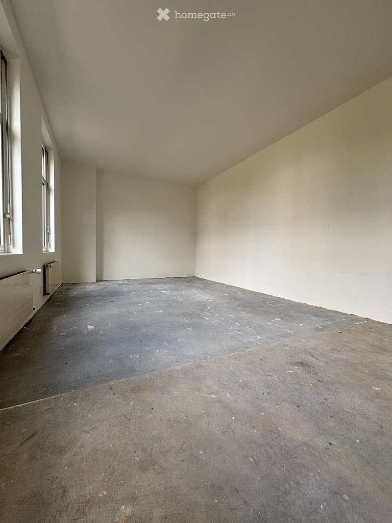 Empty room with bare walls and concrete floor, featuring a small window and a radiator on the left side.