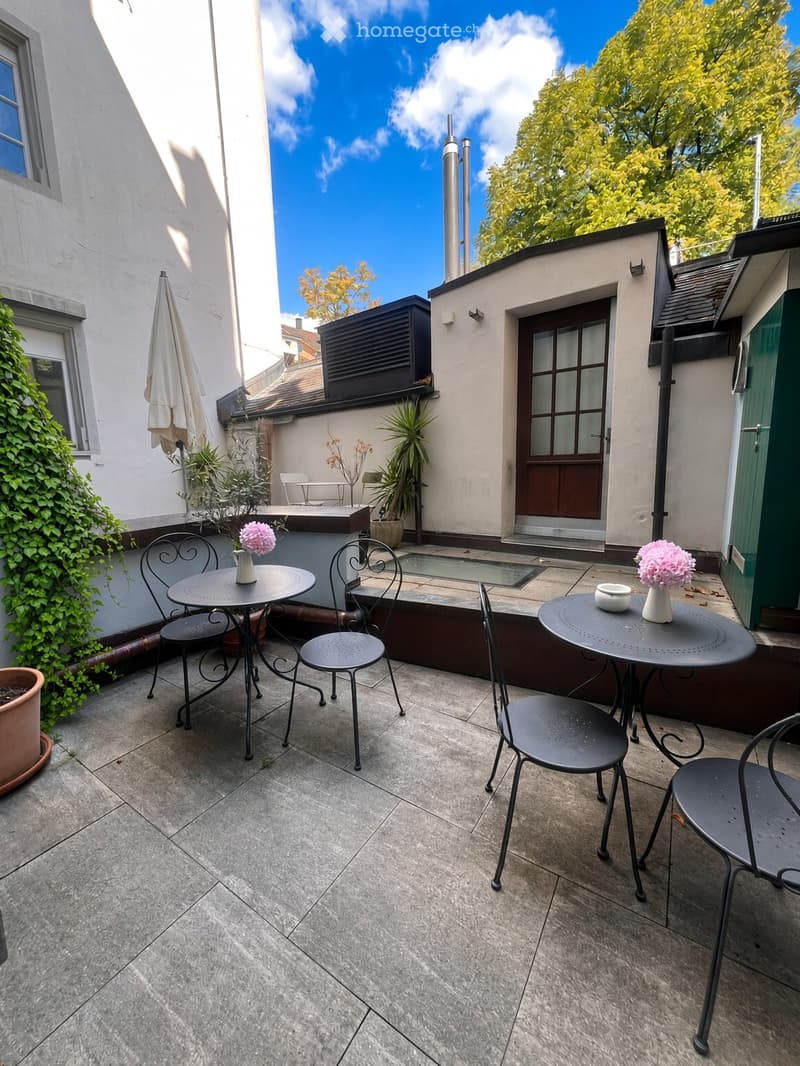A small, enclosed patio with tiled flooring, two round tables with chairs, vases with pink flowers, and an umbrella leaning against the wall.
