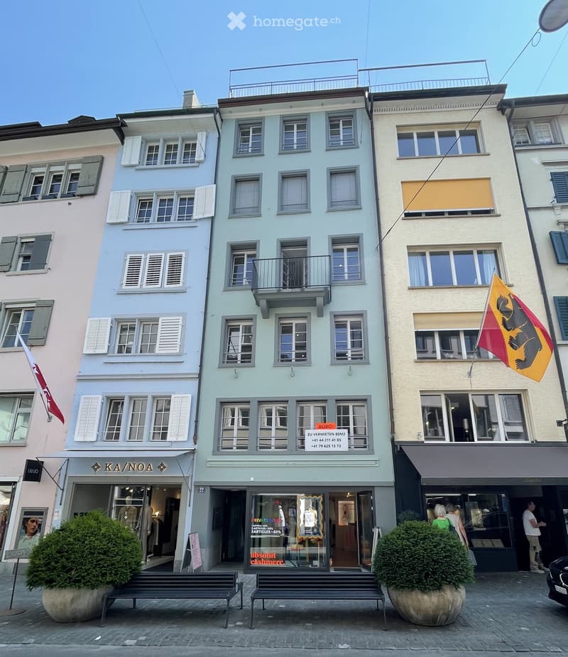 Three-story building with colorful facades, multiple windows, and a balcony on the second floor. The ground floor features storefronts with signage and mannequins in display windows.