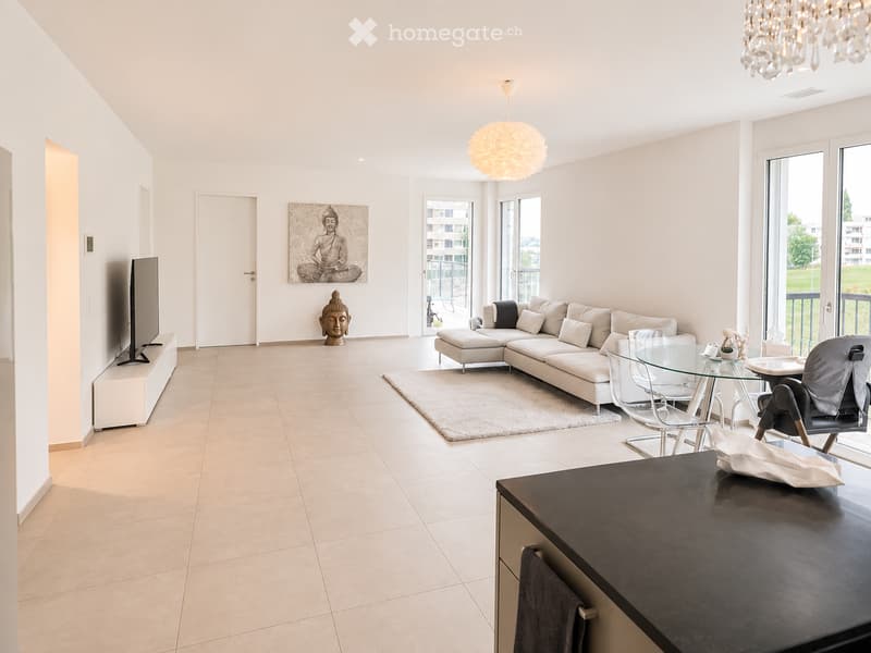 Modern living room with white walls, beige tiles, large windows, and a balcony. Features a white sectional sofa, glass coffee table, dining table with chairs, TV stand, and decorative art pieces.