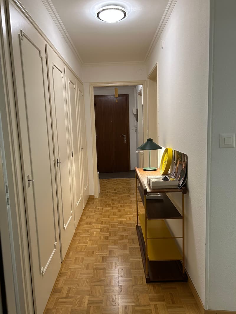 A well-lit hallway with wooden herringbone flooring, white walls, and several closed doors. A small table with a lamp and decorative items is placed against the wall.