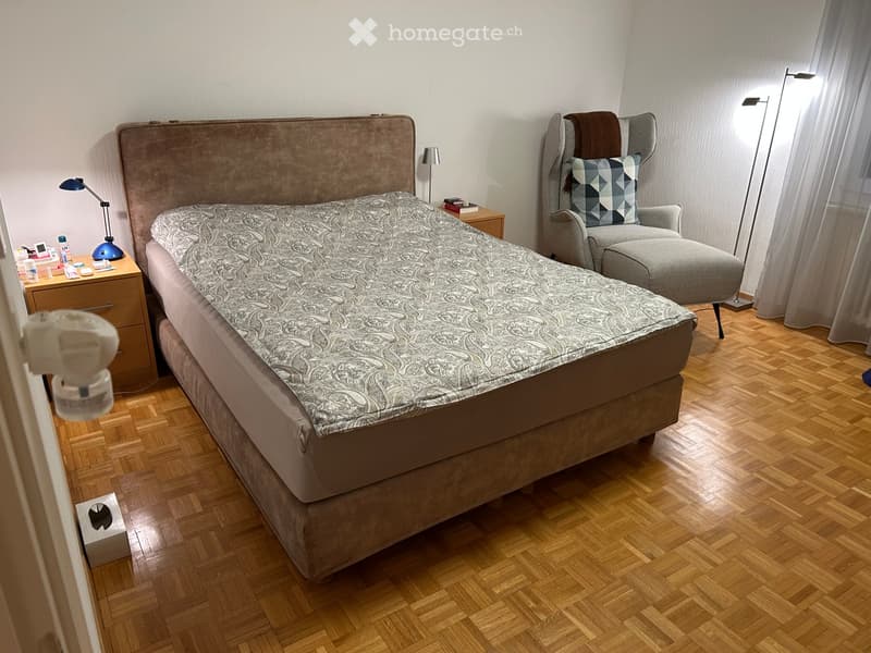 A well-furnished bedroom with a large bed, nightstand, armchair, and a lamp. The room features wooden flooring and large windows with curtains.