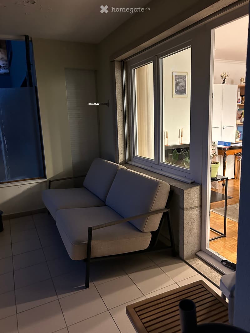 Modern living room with a grey sofa, wooden coffee table, large windows, and a view into an adjacent room with a desk and shelves.